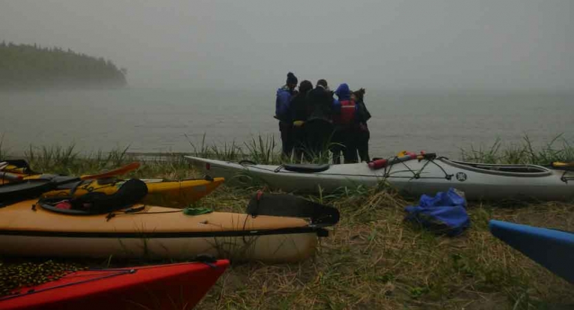 a group of outward bound students huddle in a circle on the shoreline of a lake. kayaks rest on the shore. 